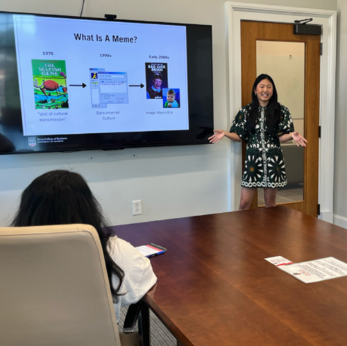 Sophia Beasley, a senior majoring in marketing with an emphasis in business analytics and digital marketing, presents her research paper, “Laugh Now, Litigate Later: Why Memes Challenge Foundations of Intellectual Property in front of large monitor in a conference room. 