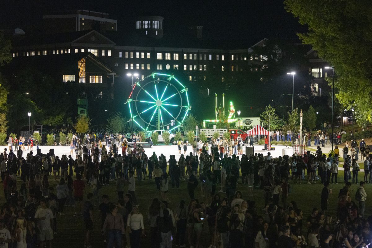 A large nighttime crowd gathers on a university campus lawn, with a brightly lit Ferris wheel and carnival rides in the background. University buildings and trees surround the event.