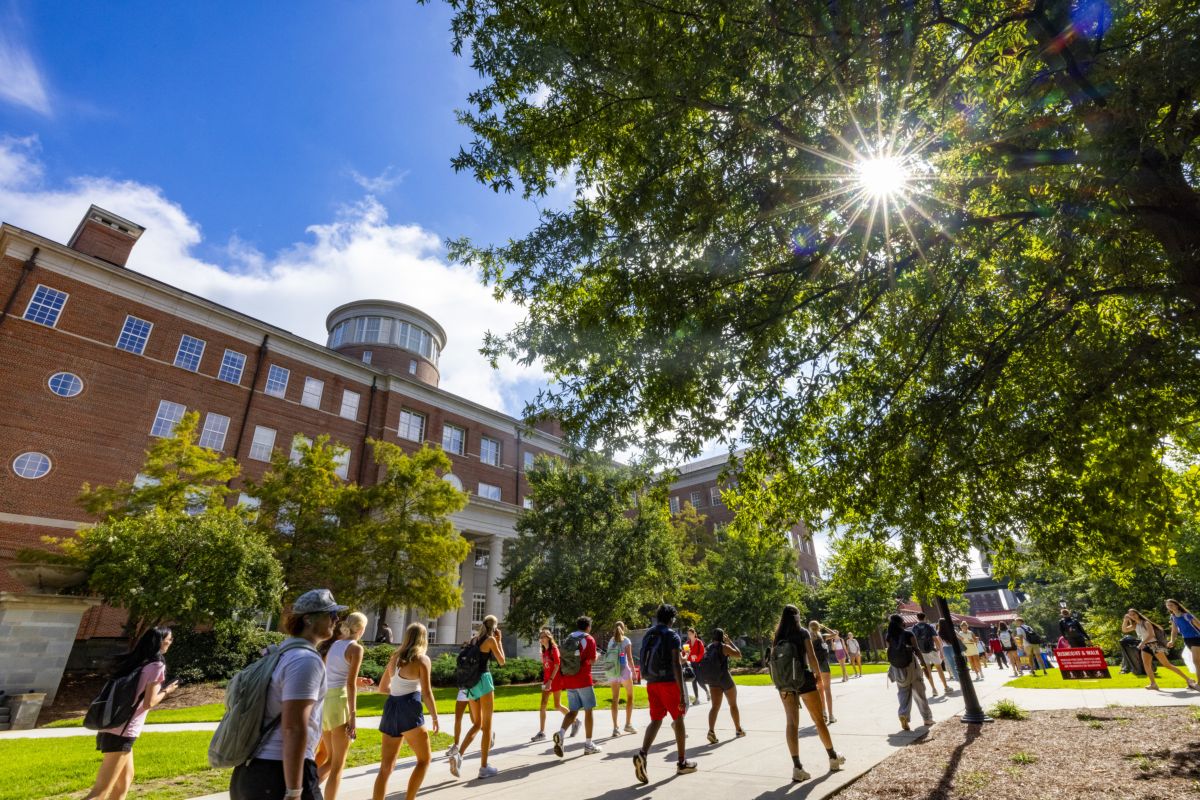 Students with backpacks walk along a sunny campus sidewalk past a large red-brick academic building with a rounded tower, framed by tree branches.