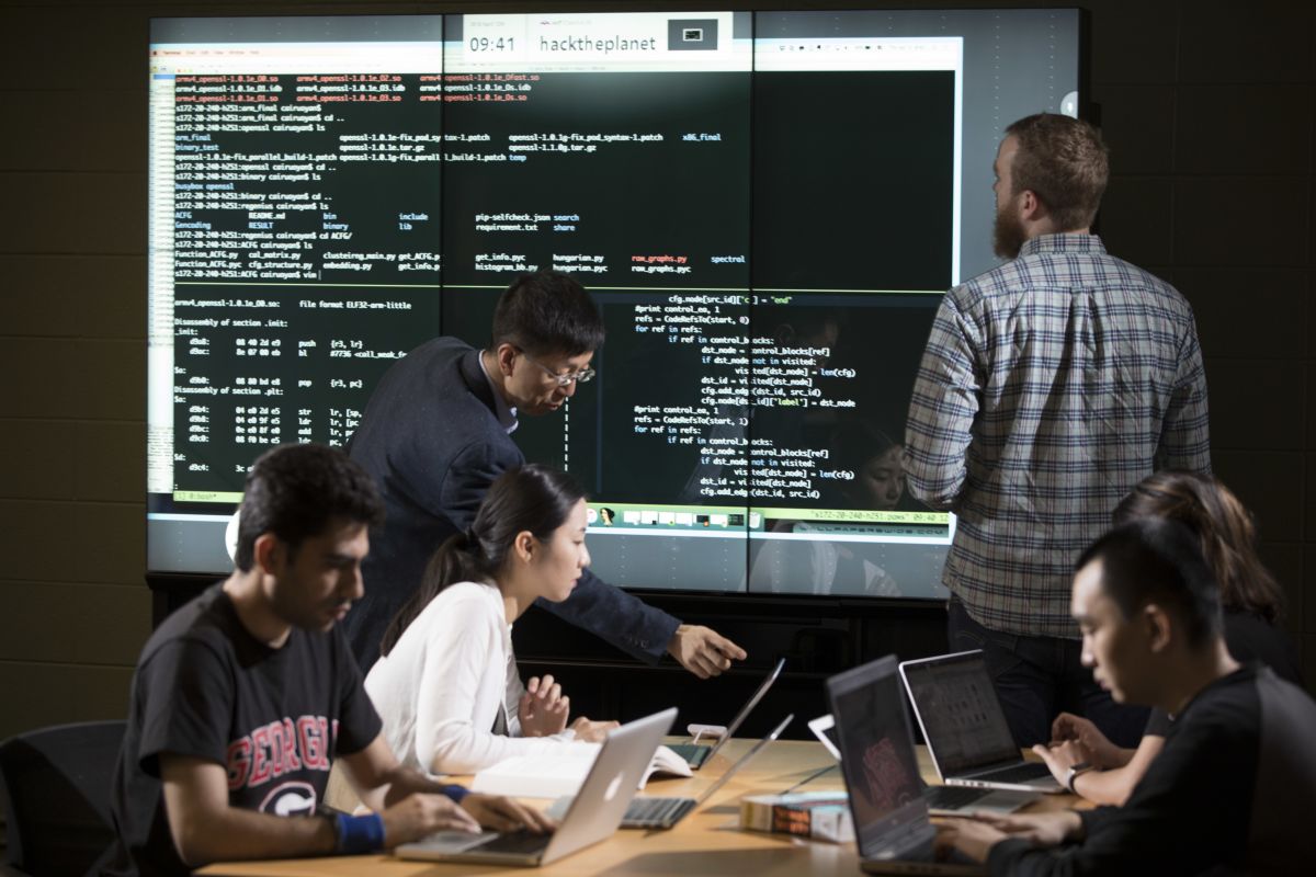 Students work on laptops at a table while an instructor and another student review lines of code on a large wall of monitors.