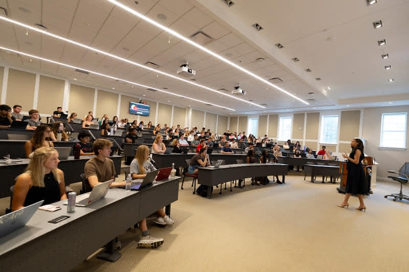 A presenter stands at the front of a brightly lit, modern lecture hall addressing rows of students seated at tiered curved desks with laptops.