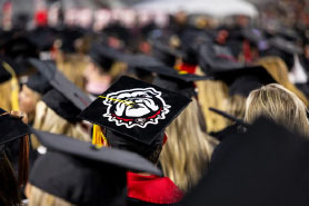 A graduate wearing a black graduation cap decorated with a bulldog logo sits amidst a large crowd of graduates in black caps and gowns.