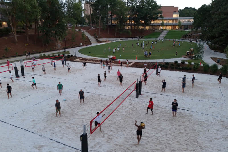 An elevated view of students playing sand volleyball across several courts adjacent to a green central lawn and campus buildings at dusk.