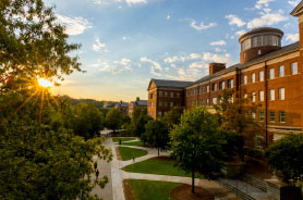 Sunlight bursts through lush green trees next to a large, multi-story brick campus building with a distinctive circular tower and paved walkways during golden hour.