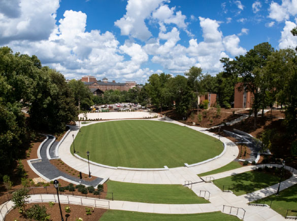 An aerial view shows an open-air amphitheater with a neatly manicured oval lawn surrounded by curved concrete walkways, green lawns, and trees under a cloudy blue sky.