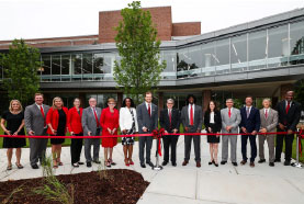 A group of university leaders and officials pose outdoors behind a long red ribbon during a ceremonial ribbon-cutting event in front of a modern campus building.